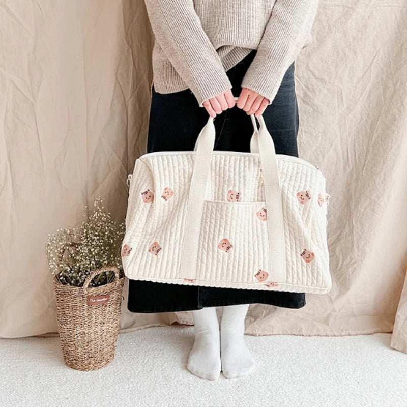 Maman tenant un sac à langer à deux mains, vue de face, du ventre aux pieds, debout à côté d’un vase de fleurs