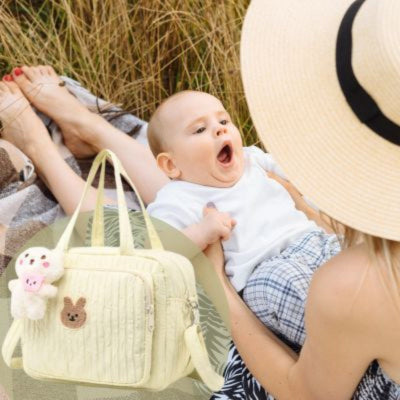 Un sac a langer lapin jaune et une maman, vue de dos, les jambes allongées, avec son bébé assis sur ses cuisses, installés sur un tapis dans un champ d’herbe