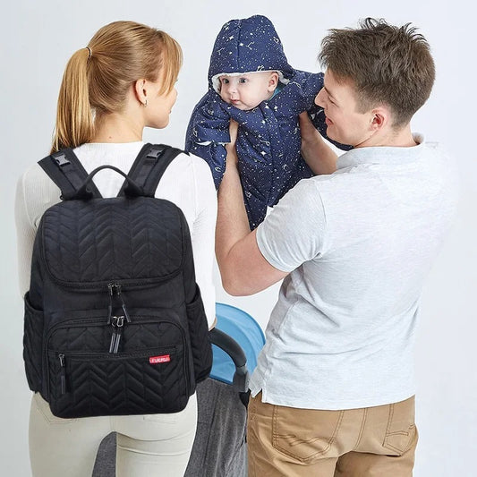 Papa avec son bébé dans les bras et maman avec un sac a langer noir sur le dos